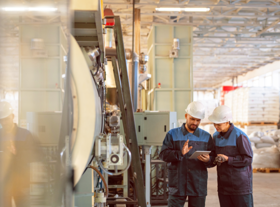Workers wearing hard hats with a tablet in an industrial facility with machinery. Workers wearing hard hats with a tablet in an industrial facility with machinery.