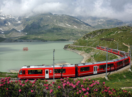 A red Rhaetian Railway train travels along a mountain lake through a blooming Alpine landscape. A red Rhaetian Railway train travels along a mountain lake through a blooming Alpine landscape.