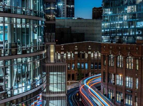 Illuminated railway line at night, modern/historic buildings, light trails of a train.