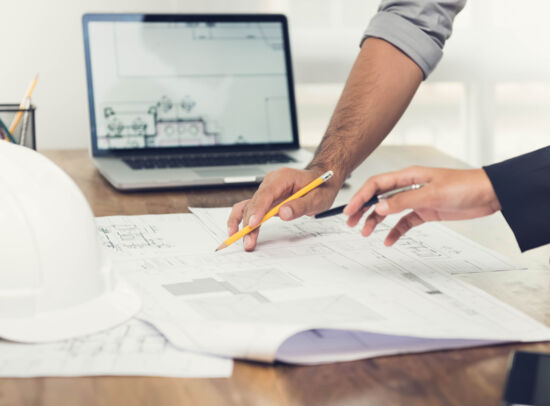 Two people working on construction plans at a table, with a laptop and a white safety helmet.