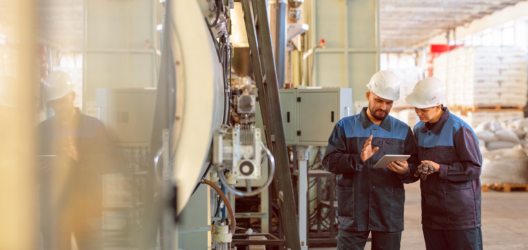 Workers wearing hard hats with a tablet in an industrial facility with machinery. Workers wearing hard hats with a tablet in an industrial facility with machinery.