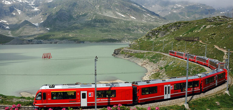A red Rhaetian Railway train travels along a mountain lake through a blooming Alpine landscape. A red Rhaetian Railway train travels along a mountain lake through a blooming Alpine landscape.