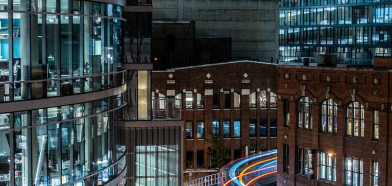 Illuminated railway line at night, modern/historic buildings, light trails of a train.