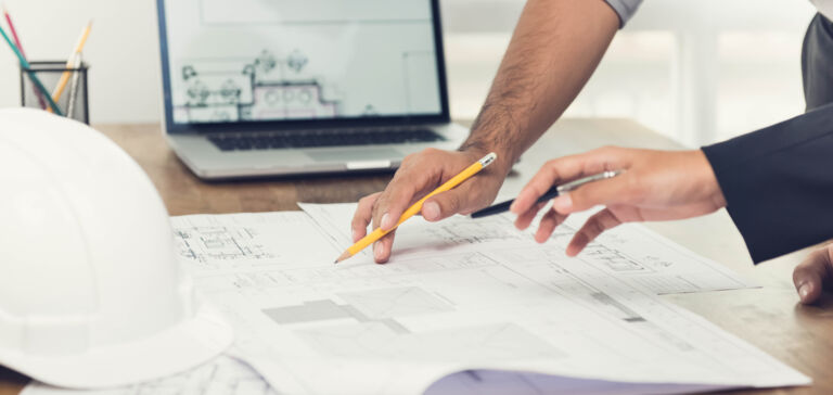 Two people working on construction plans at a table, with a laptop and a white safety helmet.
