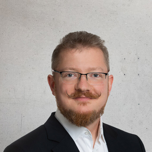 Portrait photo of Andreas Wenz, a man with glasses, a beard and a moustache, against a grey background.